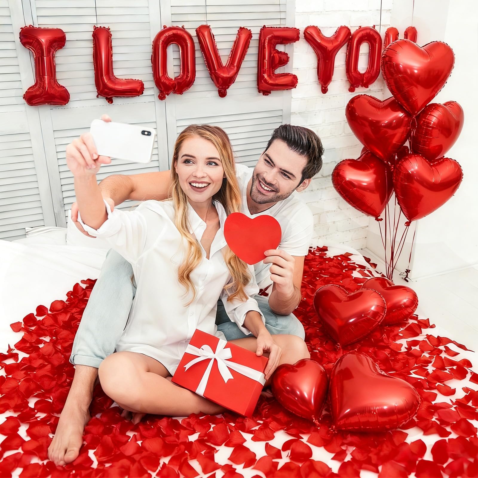 Couple taking a selfie surrounded by red heart balloons and rose petals, with 'I LOVE YOU' balloons in the background.