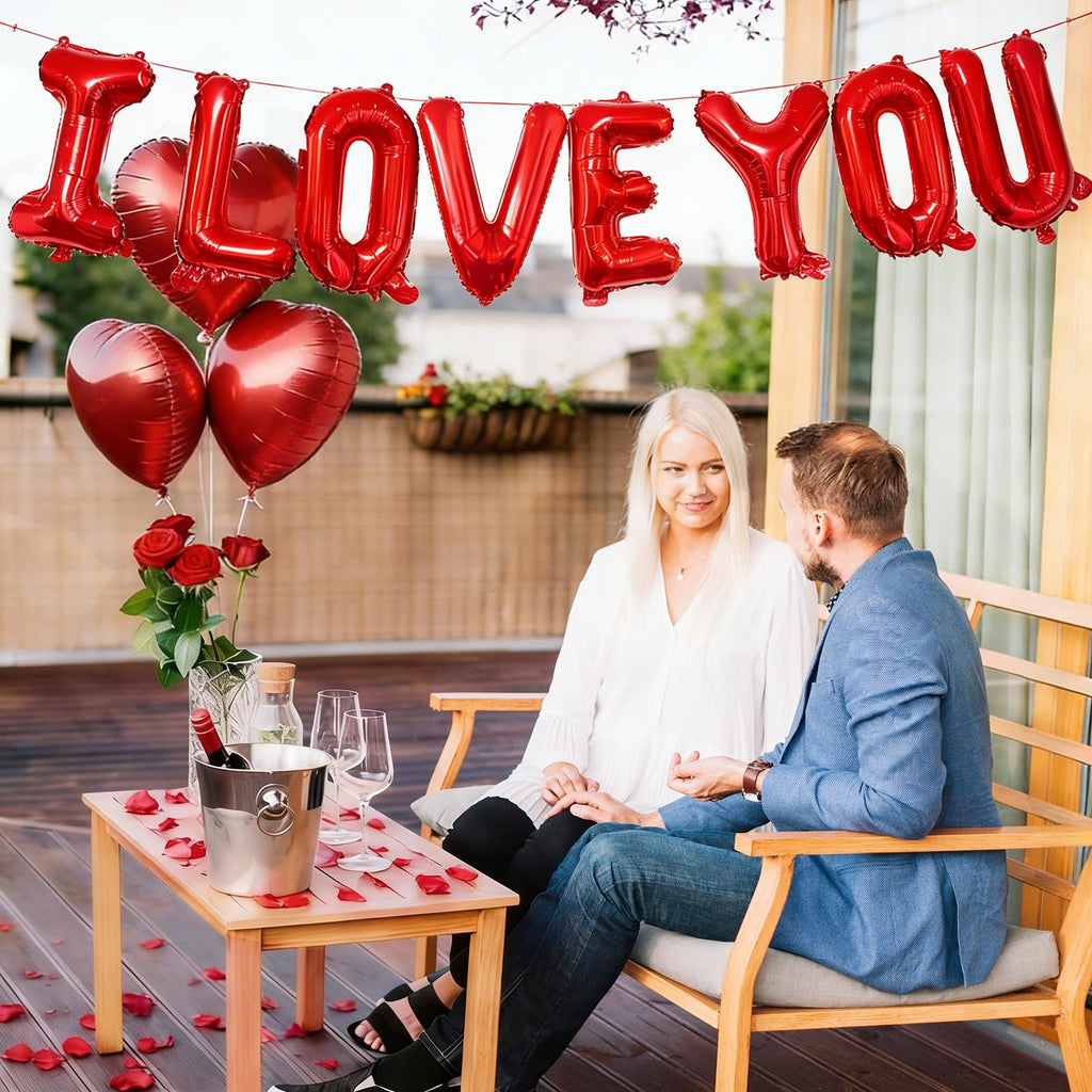 Couple on a balcony with red heart and 'I LOVE YOU' foil balloons, wine, and roses