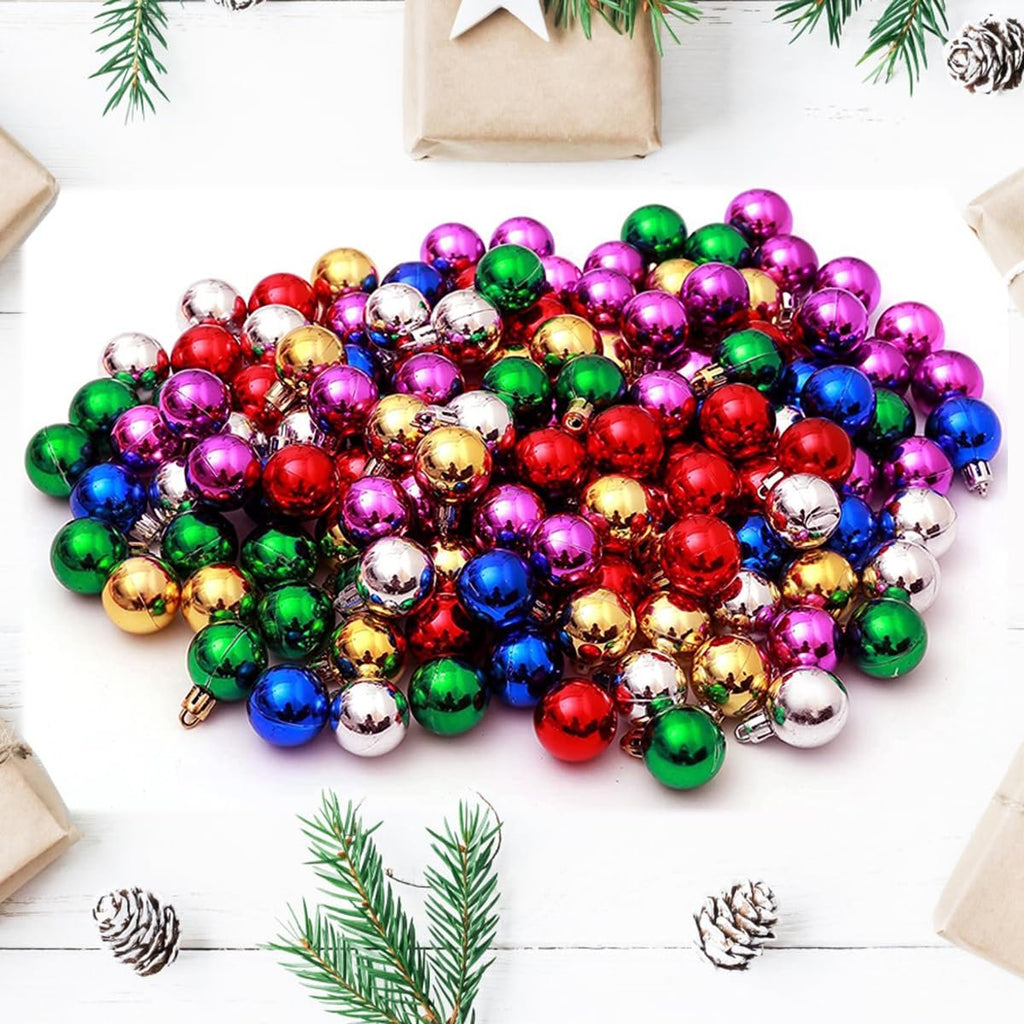 Pile of small, colorful Christmas ball ornaments in red, green, gold, blue, purple, and silver, arranged on a white surface with pine sprigs and gift boxes.