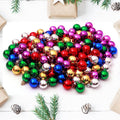 Pile of small, colorful Christmas ball ornaments in red, green, gold, blue, purple, and silver, arranged on a white surface with pine sprigs and gift boxes.