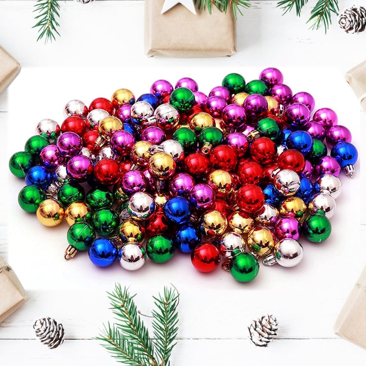 Pile of small, colorful Christmas ball ornaments in red, green, gold, blue, purple, and silver, arranged on a white surface with pine sprigs and gift boxes.