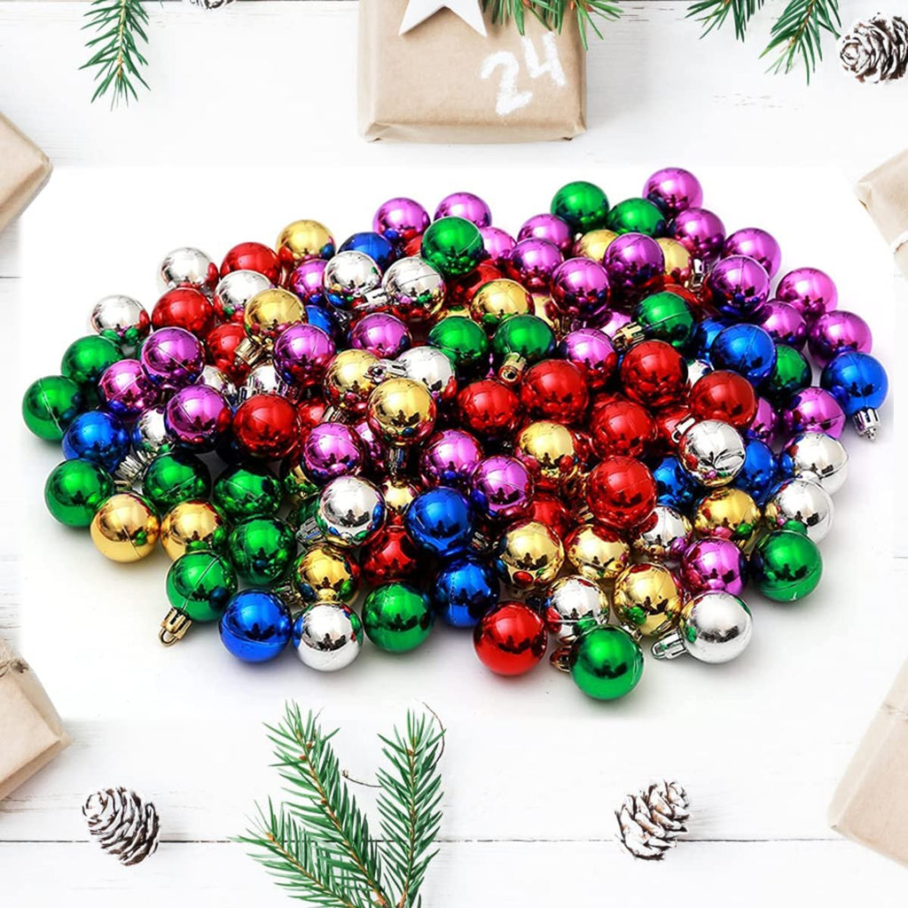 Pile of small, colorful Christmas ball ornaments in red, green, gold, blue, purple, and silver, scattered on a white surface with pine sprigs and gift boxes.