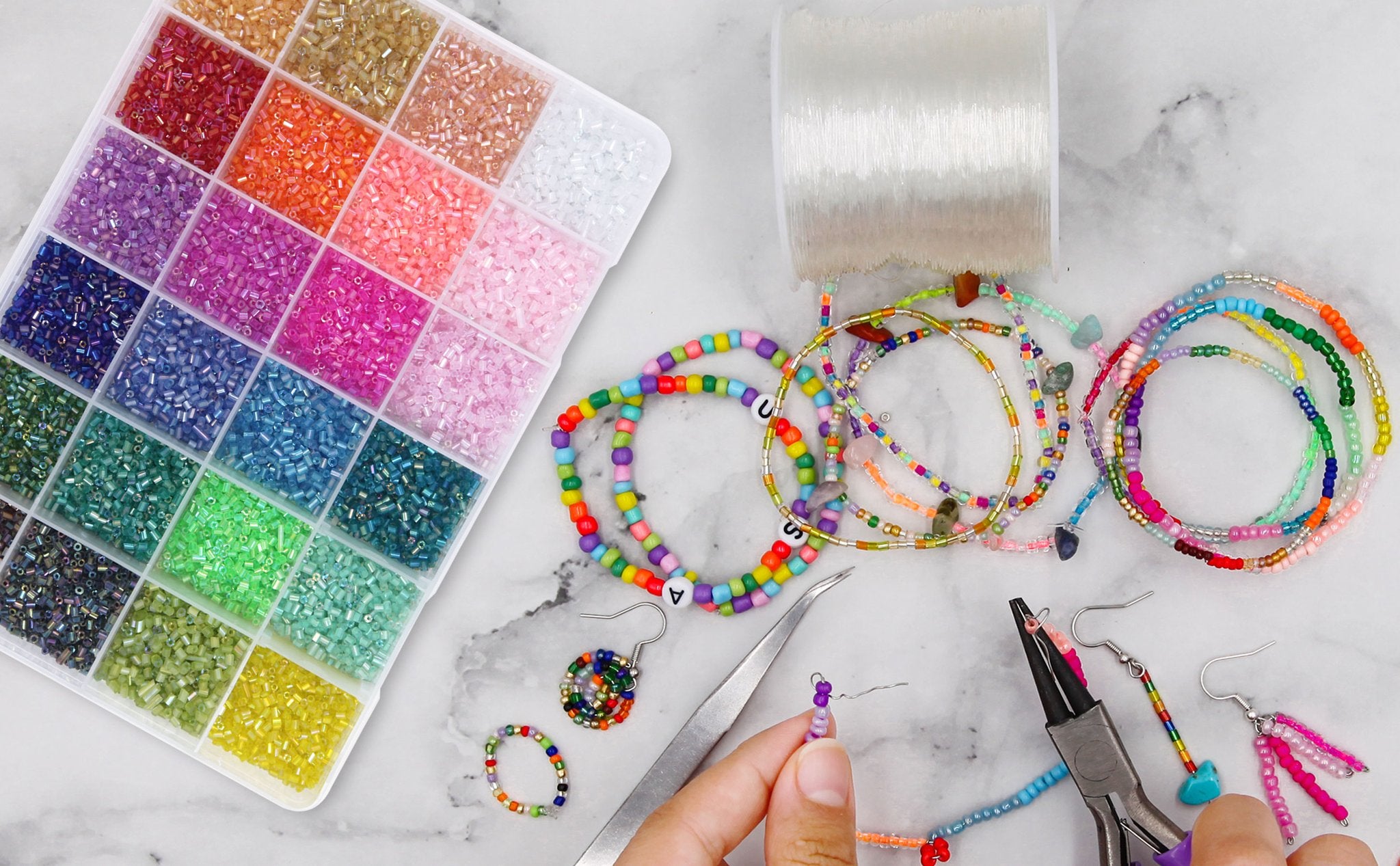 Flat lay of a bead kit with 24 compartments of colorful seed beads, spools of elastic string, pliers, tweezers, and several finished bracelets and earrings.