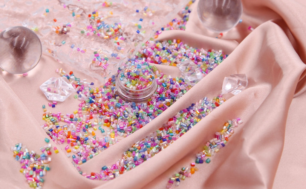 Close-up of colorful bugle glass seed beads scattered on pink satin fabric with clear acrylic decorative elements.