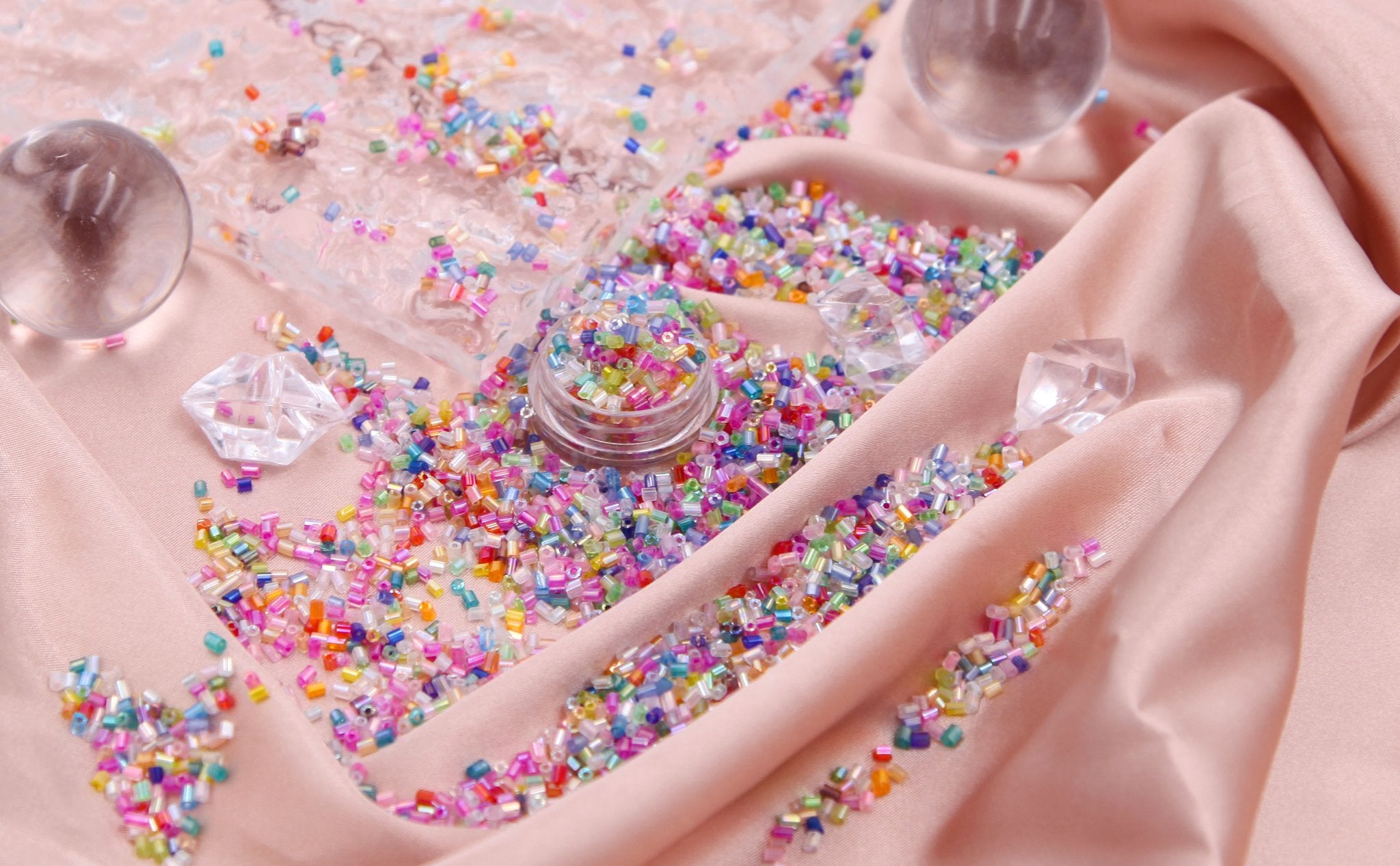 Close-up of colorful bugle glass seed beads scattered on pink satin fabric with clear acrylic decorative elements.