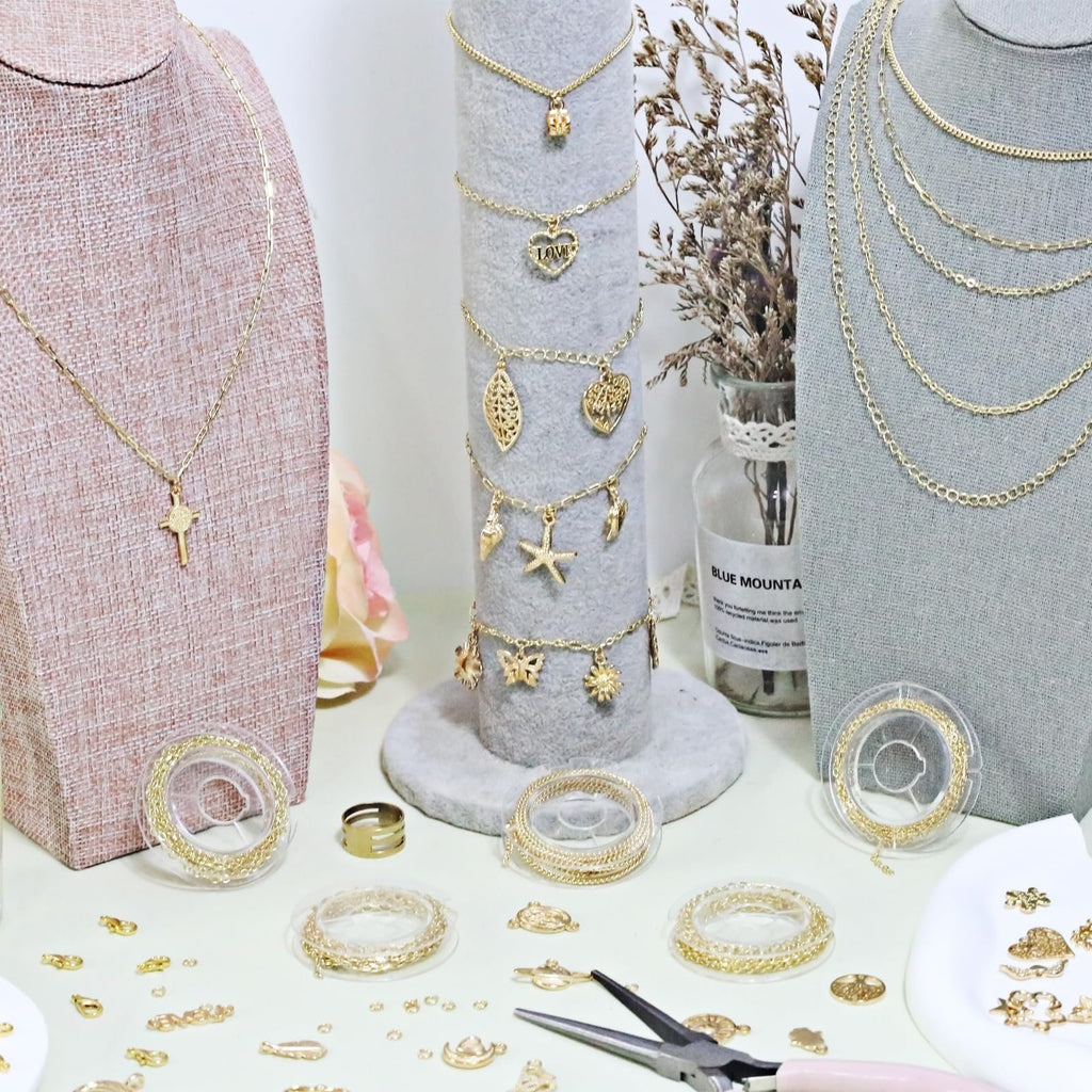 Gold jewelry making kit components including chains, charms, clasps, jump rings, and pliers, displayed on a table with jewelry busts.