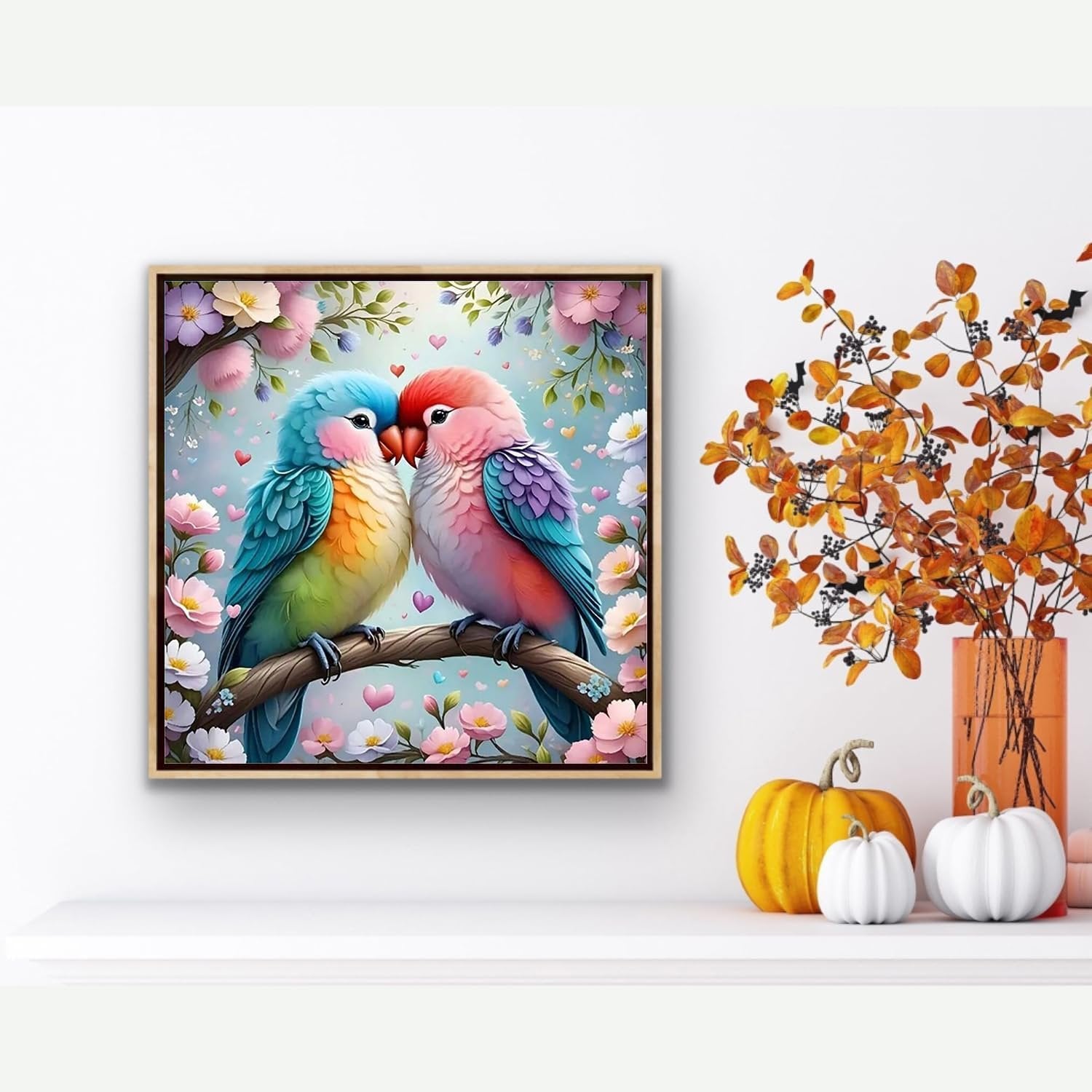Two colorful lovebirds perched on a branch with flowers and hearts, framed art on a shelf with pumpkins and fall foliage.