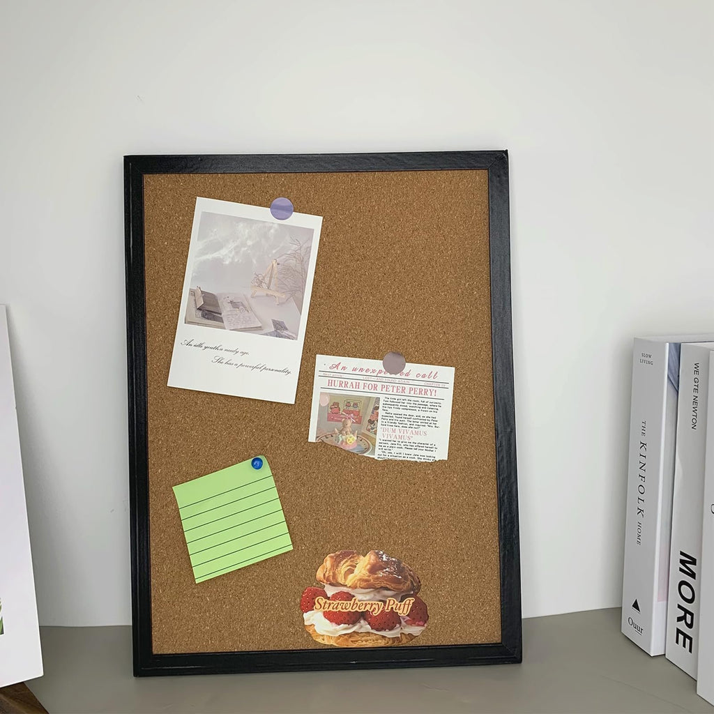 Black cork bulletin board with oak wood finish frame, decorated with photos and notes, standing next to books.