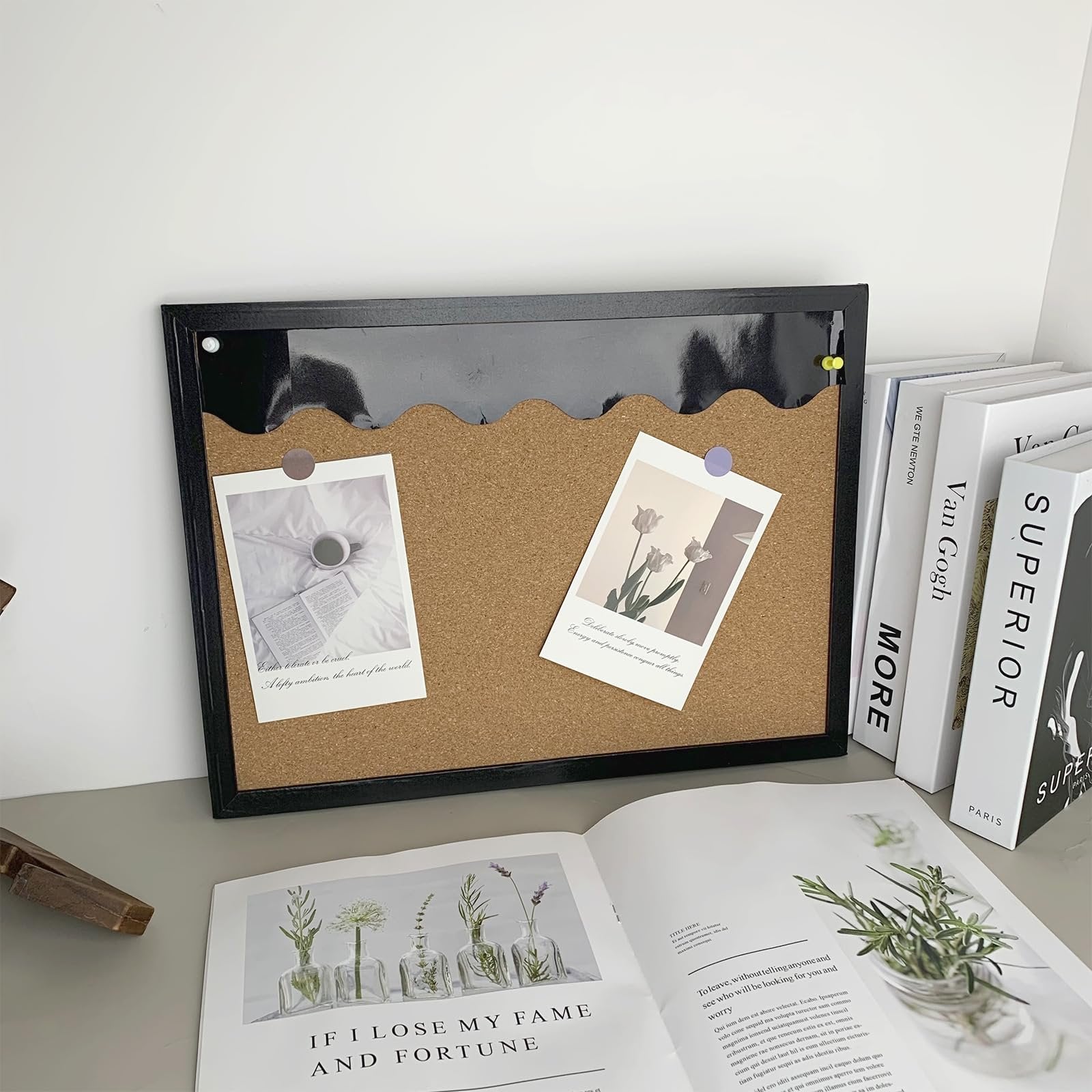 Black cork bulletin board with scalloped edge and two photos pinned, displayed on a desk with books and an open magazine.
