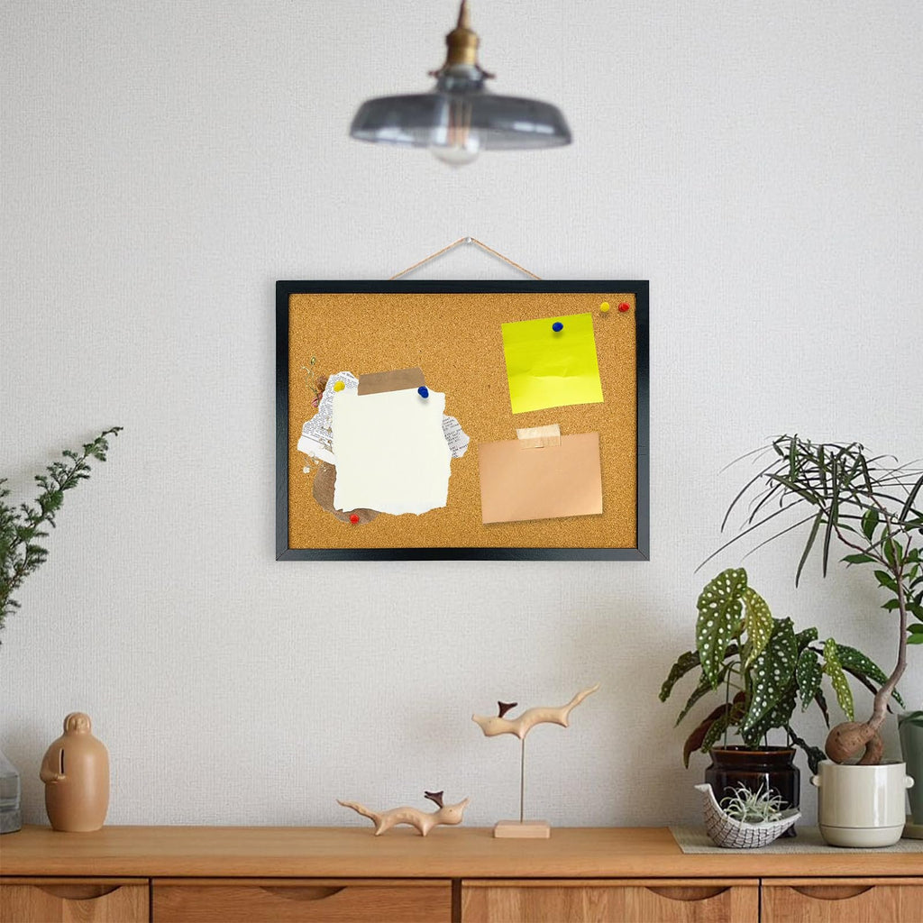 Black framed cork bulletin board with pinned papers and sticky notes, hanging on a wall above a wooden dresser with plants.