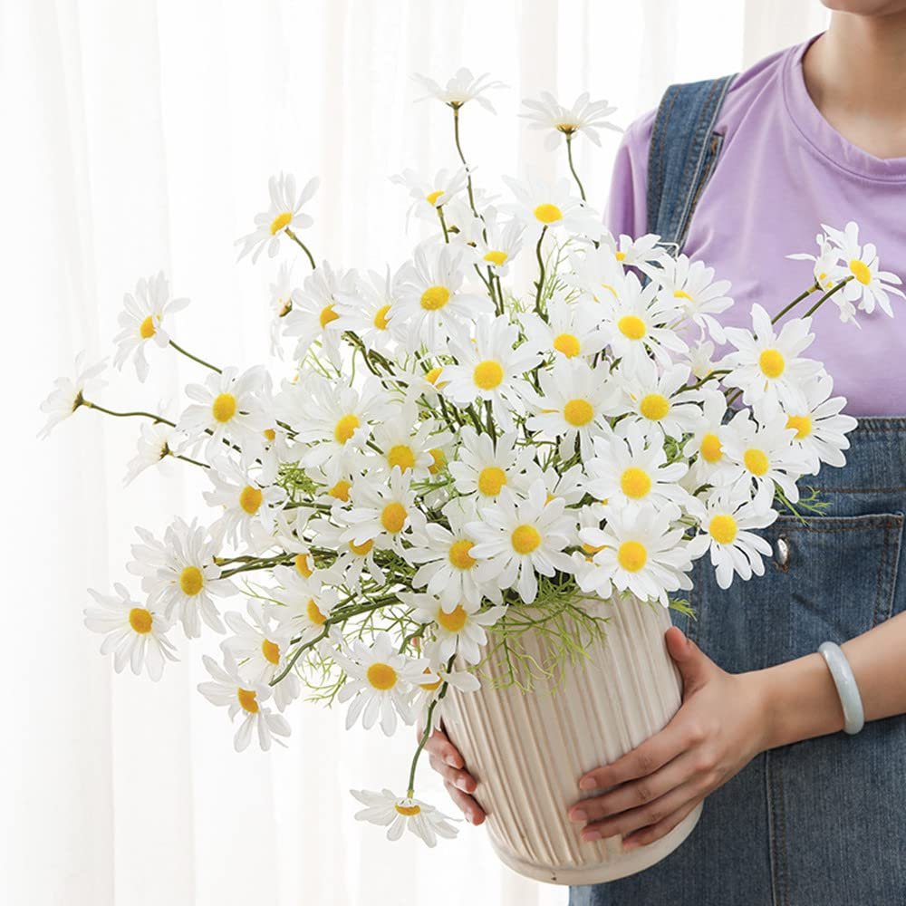 Bundle of artificial white daisy flowers in a cream vase held by a person in denim overalls