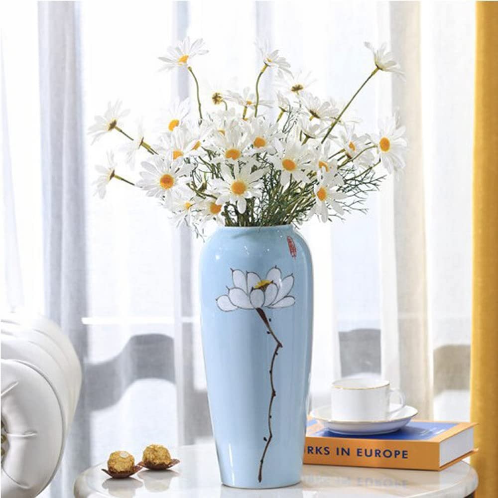 Light blue ceramic vase with white daisy flowers and a lotus design, on a table with a book and teacup.