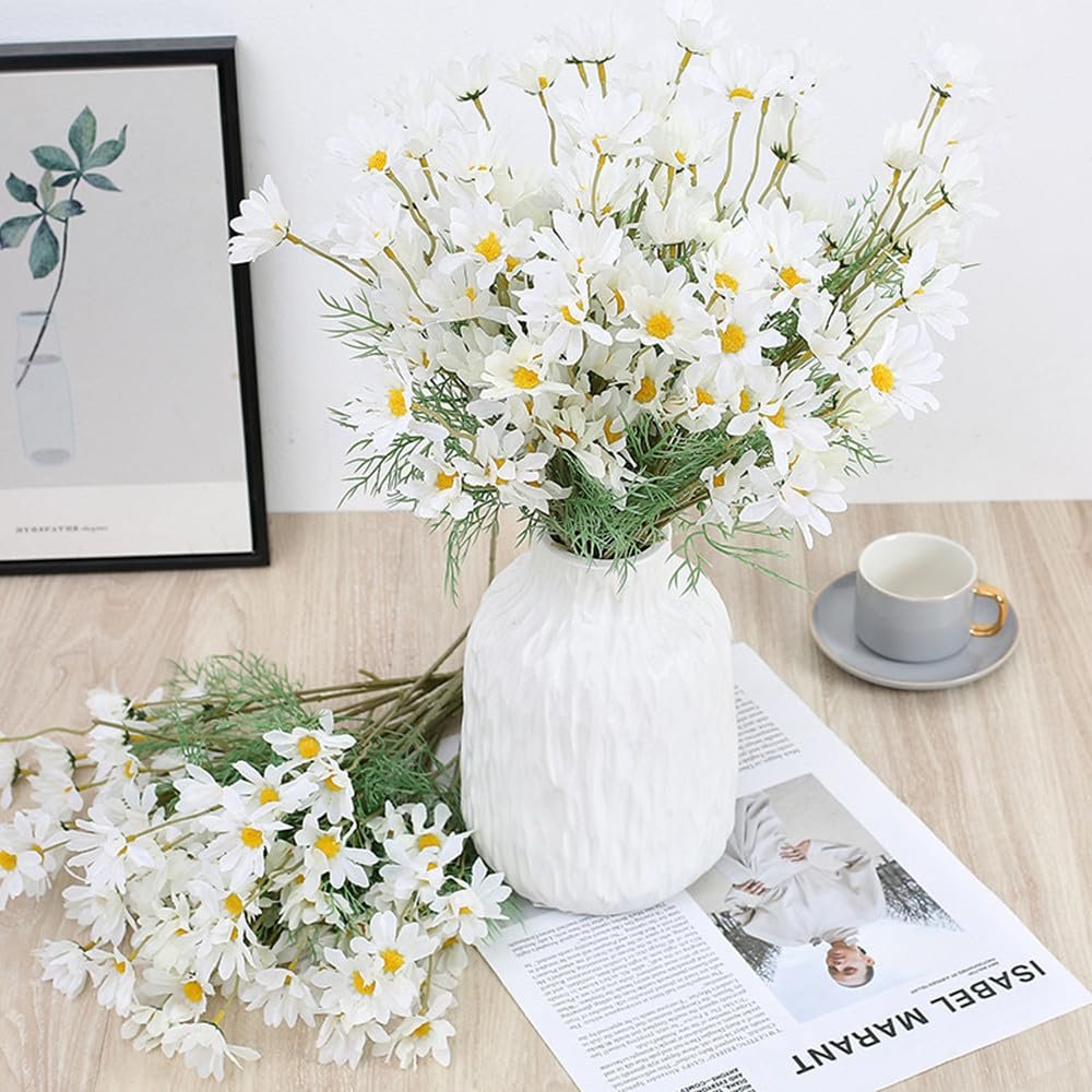 White daisy flowers in a textured white vase on a wooden surface with a framed picture and a coffee cup.