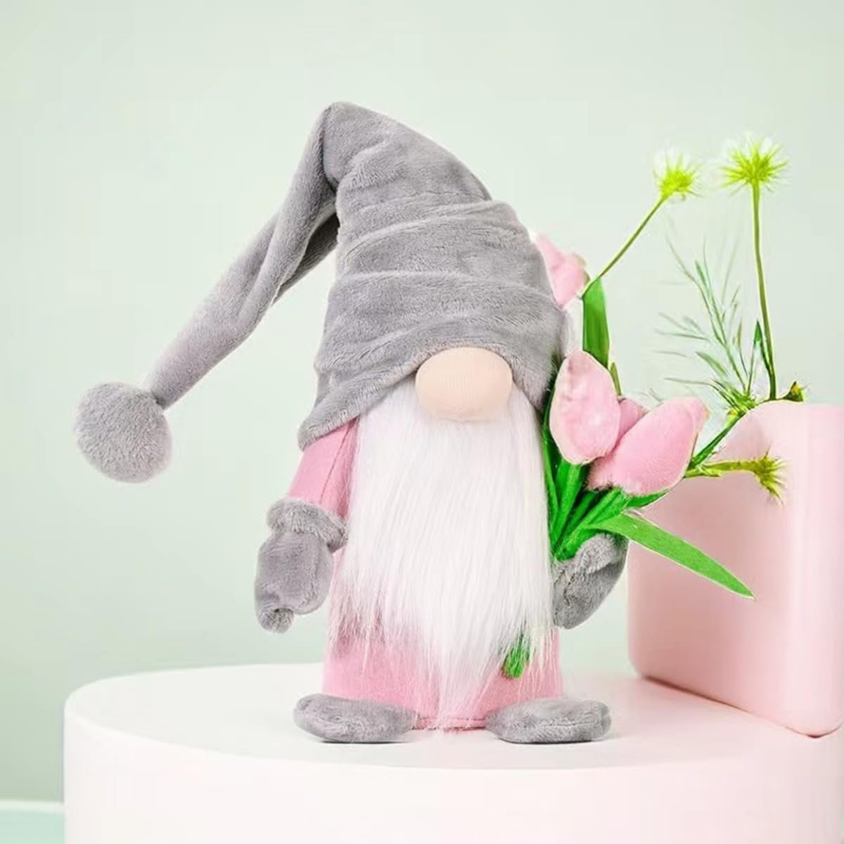 Plush gnome doll with a grey hat and pink outfit holding a bouquet of pink tulips, set against a light green background.