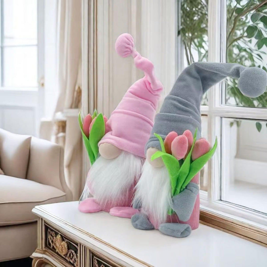 Two plush gnome dolls with tulips, one pink and one grey, sitting on a table by a window.