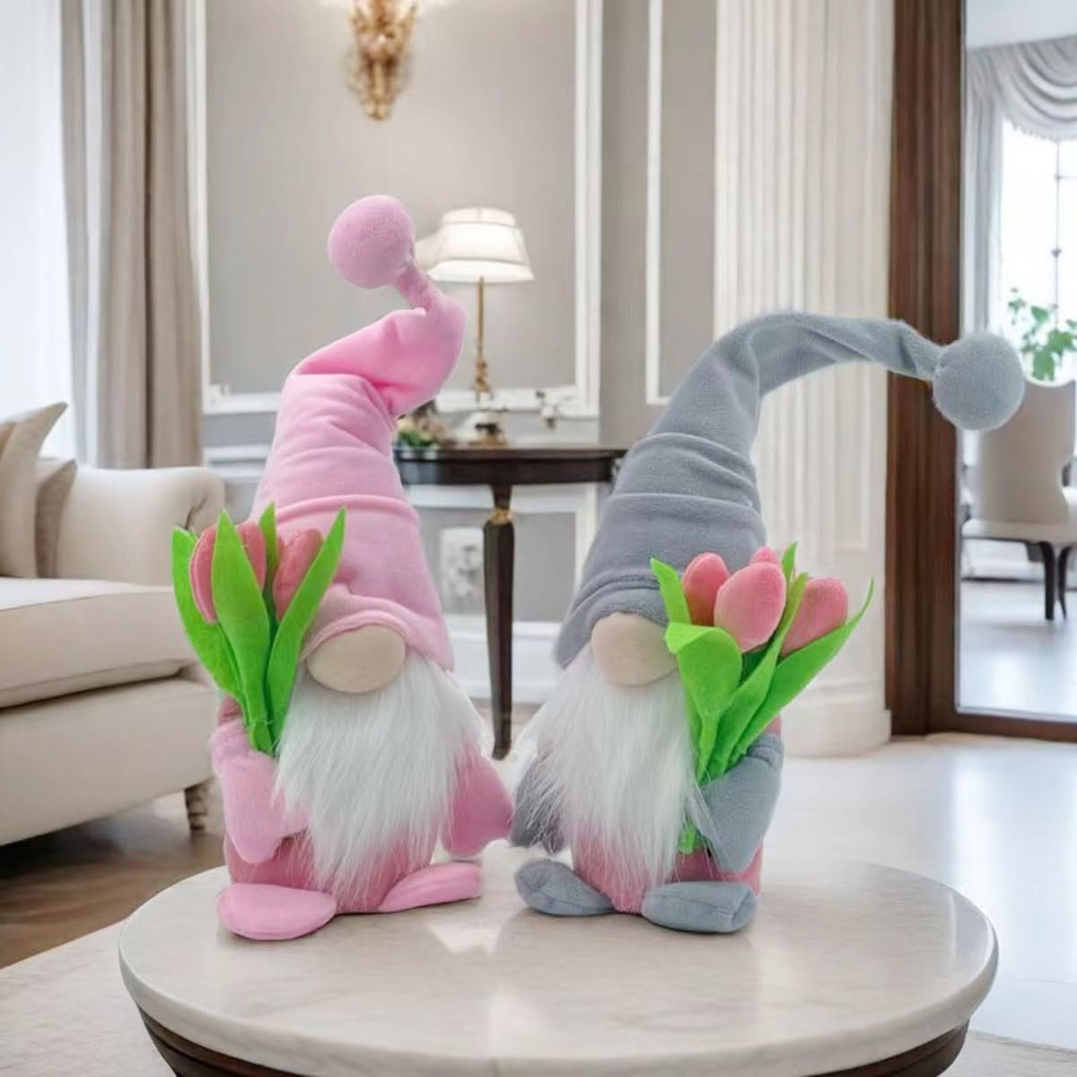 Two plush gnome dolls with tulips, one pink and one grey, sitting on a marble table in a living room.
