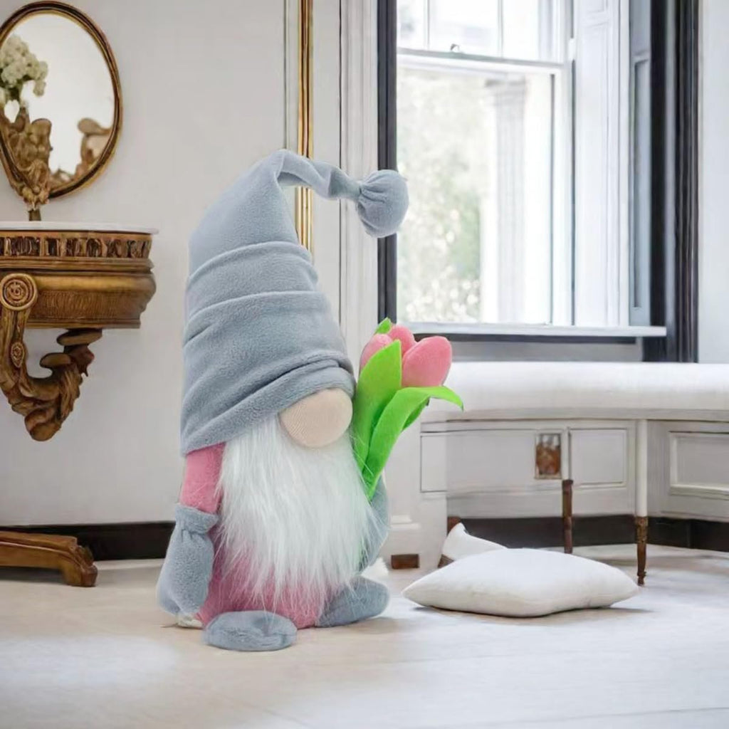 Grey and pink gnome plush doll holding pink tulips, seated indoors on a light wood floor.