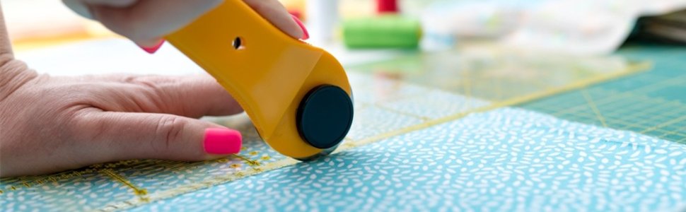 Close-up of a hand using a yellow rotary cutter to cut blue patterned fabric on a green cutting mat.