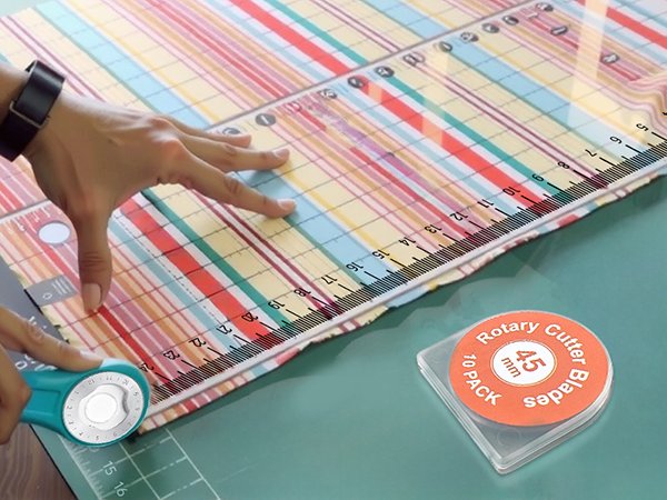 Hands using a rotary cutter and ruler to cut patterned fabric on a cutting mat; container of blades nearby.