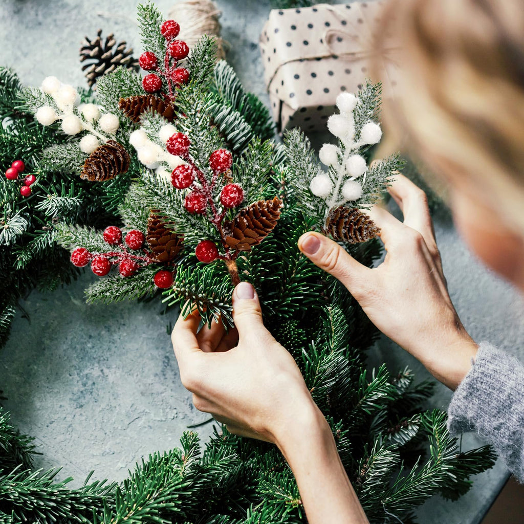 Hands arranging artificial pine branches with red berries, white frosted berries, pinecones, and hemp bows on a grey surface.