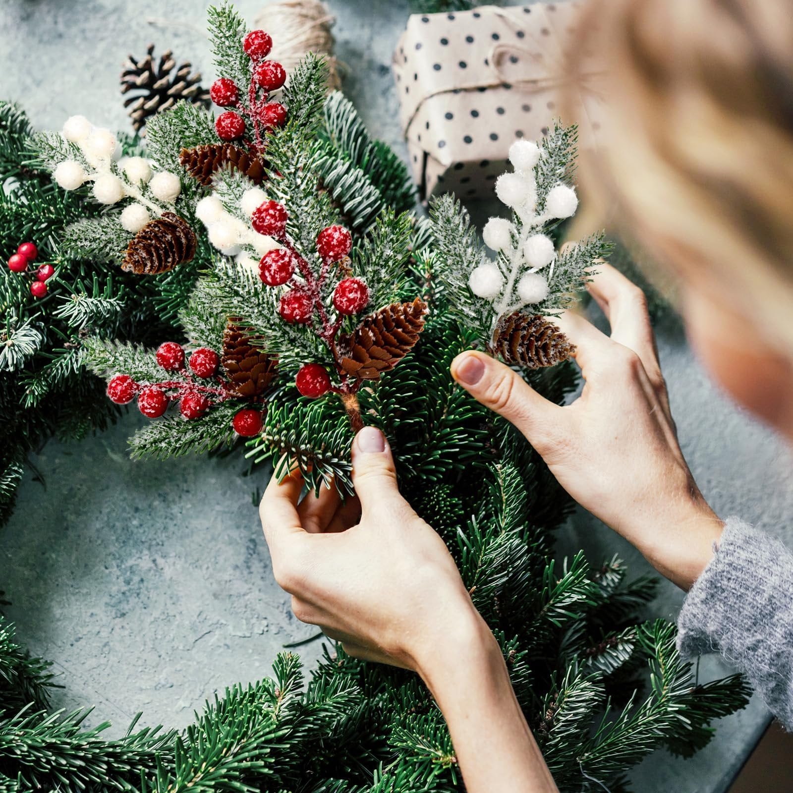 Hands arranging artificial pine branches with red berries, white frosted berries, pinecones, and hemp bows on a grey surface.