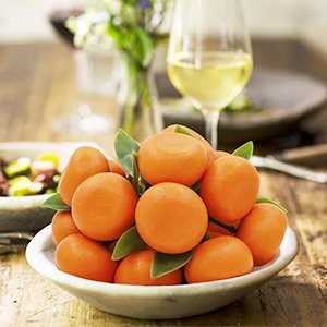 Bowl of 15 mini artificial oranges with green leaves, set on a wooden table with a glass of white wine and a vase of flowers.