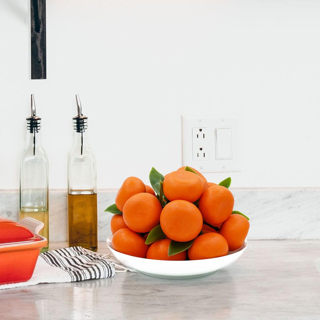 Bowl of mini artificial oranges with green leaves on a kitchen counter next to oil bottles.