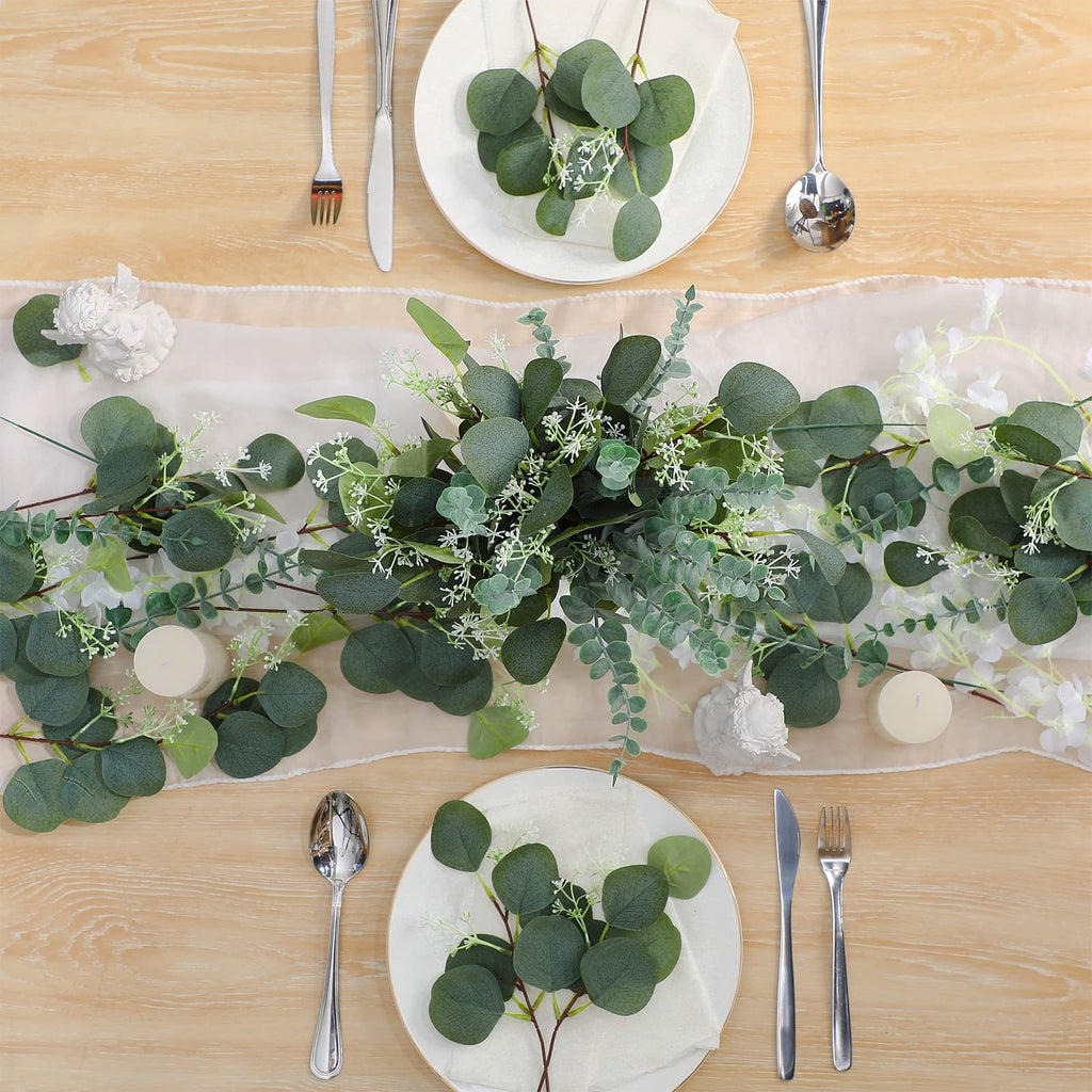 Table setting with artificial eucalyptus garland and white flowers, plates, cutlery, and candles.