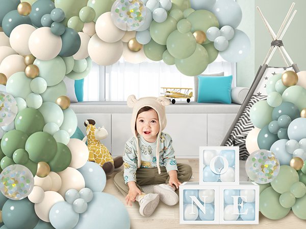 Baby sitting in front of a dusty blue green balloon garland arch kit with gold confetti and decorative 'ONE' blocks