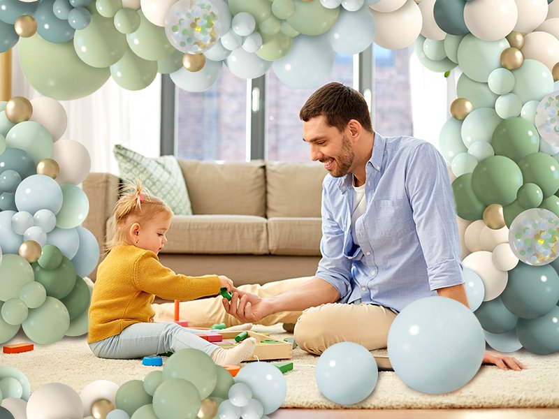 Father and daughter playing on floor surrounded by dusty blue, green, and gold balloons