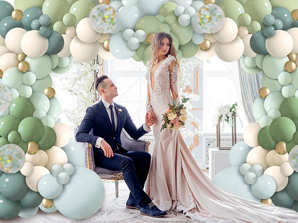 Bride and groom posing amidst a dusty blue, green, and gold balloon garland arch