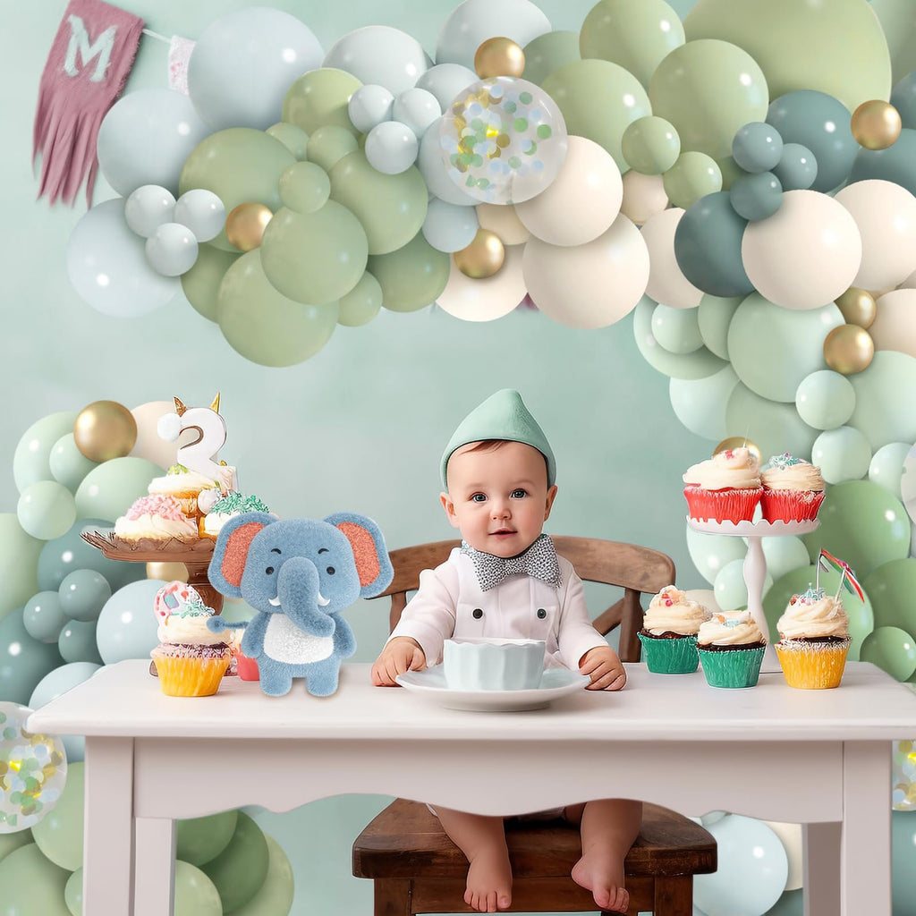 Baby sitting at a table with cupcakes and a stuffed elephant, surrounded by a balloon garland.