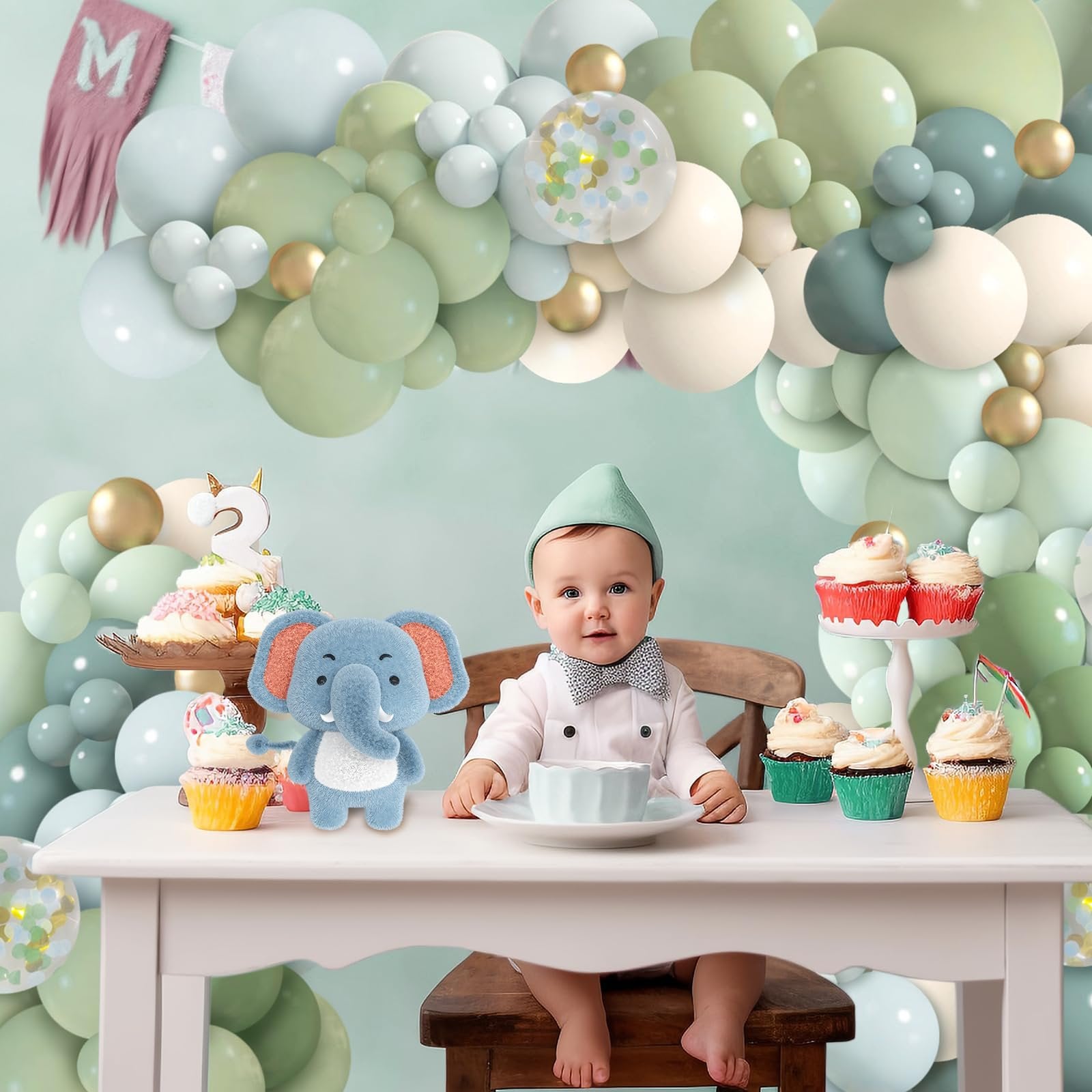 Baby sitting at a table with cupcakes and a stuffed elephant, surrounded by a balloon garland.
