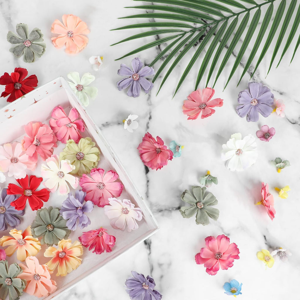 Assorted faux fabric flowers in various pastel colors scattered on a marble surface with a green palm leaf accent.