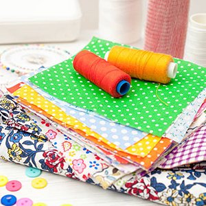 Stack of pre-cut fabric squares in various patterns with spools of thread and buttons, arranged on a white surface.