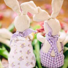 Two stuffed bunny rabbits wearing scarves and patterned outfits, surrounded by green leaves and pink petals.