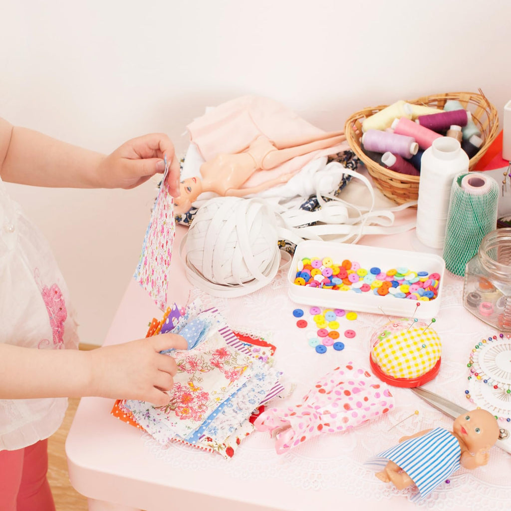 Child's hands sorting through colorful fabric squares on a pink table with sewing supplies and a doll.
