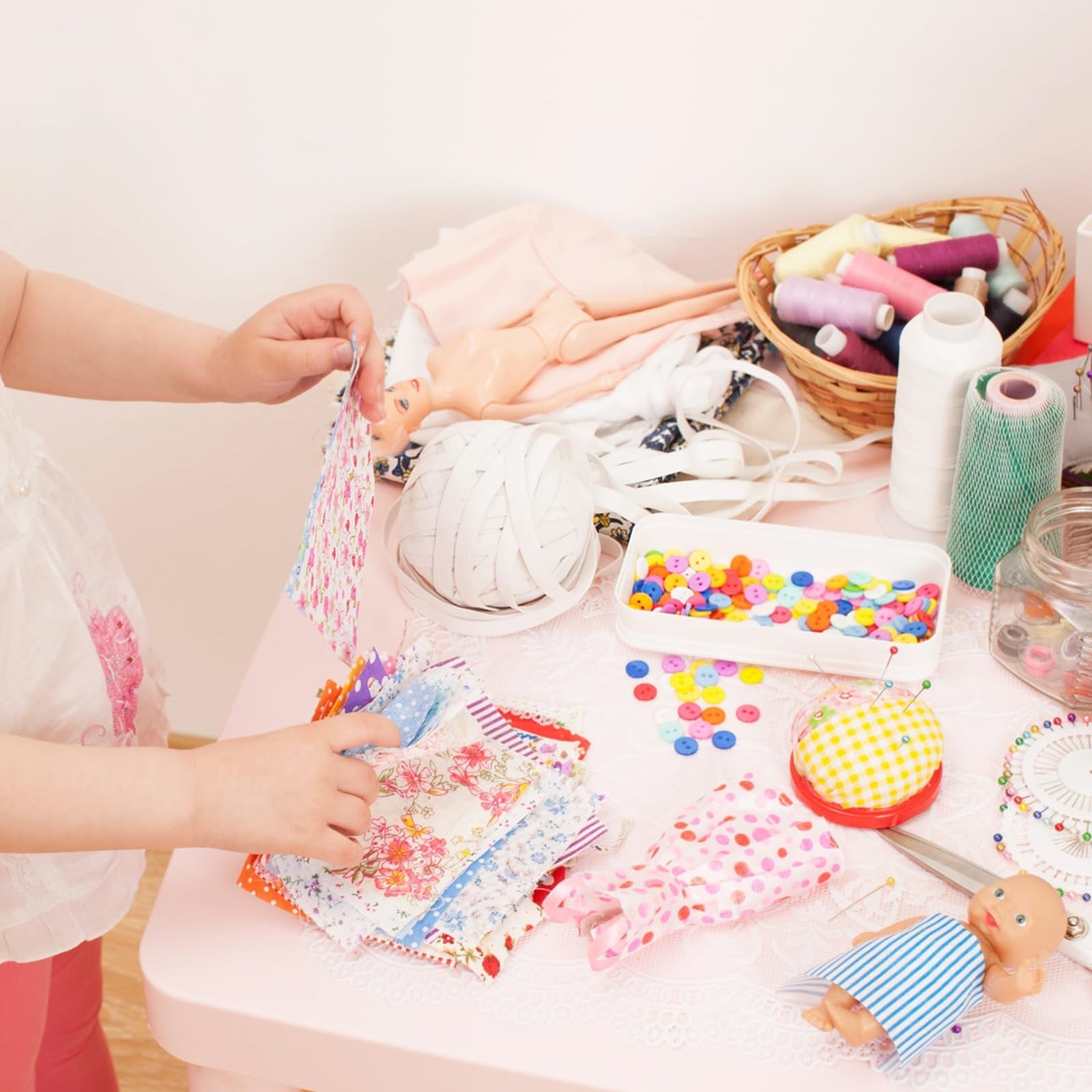Child's hands sorting through colorful fabric squares on a pink table with sewing supplies and a doll.