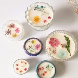 Assortment of five homemade candles decorated with dried pressed flowers and botanicals in various glass and ceramic containers.