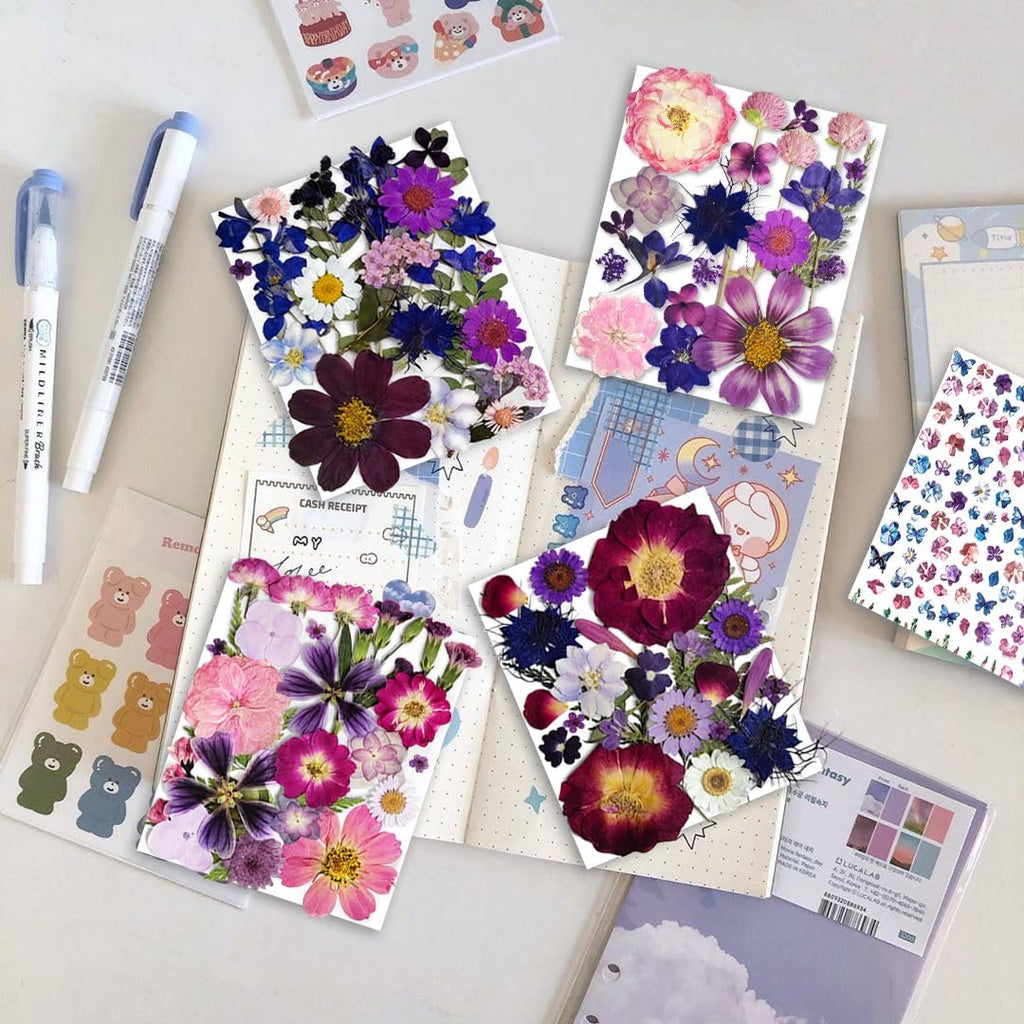 Assortment of dried pressed flowers in purple, pink, and blue hues arranged on paper, with pens and stickers nearby.