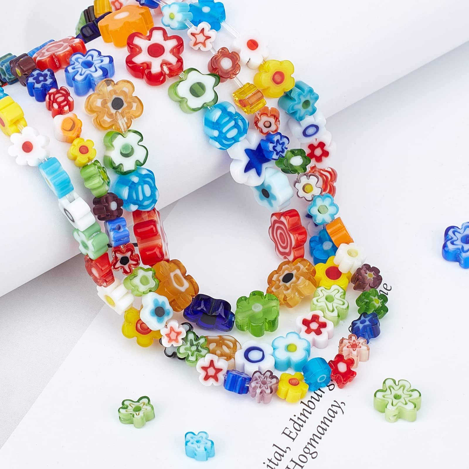 Assorted colorful millefiori glass beads in flower and star shapes, arranged in strands and scattered on a white surface.