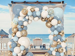 Dusty blue, white, and gold balloon arch decoration set up on a beach with ocean and chairs in the background.