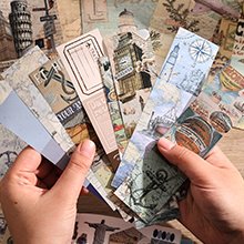 Hands holding a fan of travel-themed scrapbook paper strips with maps, landmarks, and nautical designs.