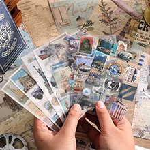 Hands holding a fan of travel-themed stickers and journaling papers, with a notebook in the background.