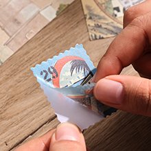 Hands peeling backing off a travel-themed sticker designed to look like a postage stamp.