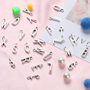 Antique silver alphabet letter charms and jump rings scattered on a pink surface with decorative pearls and pom-poms.