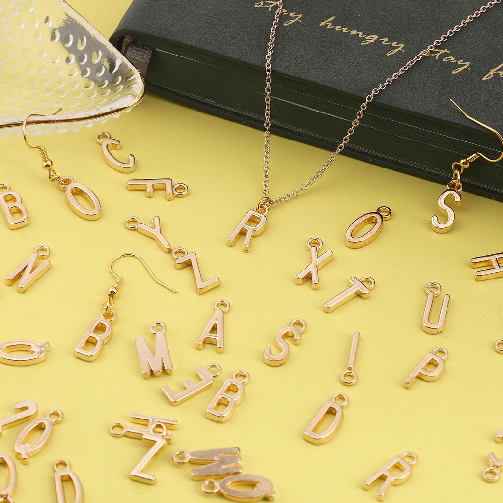 Gold alphabet letter charms and earrings scattered on a yellow background with a green book and decorative tray.