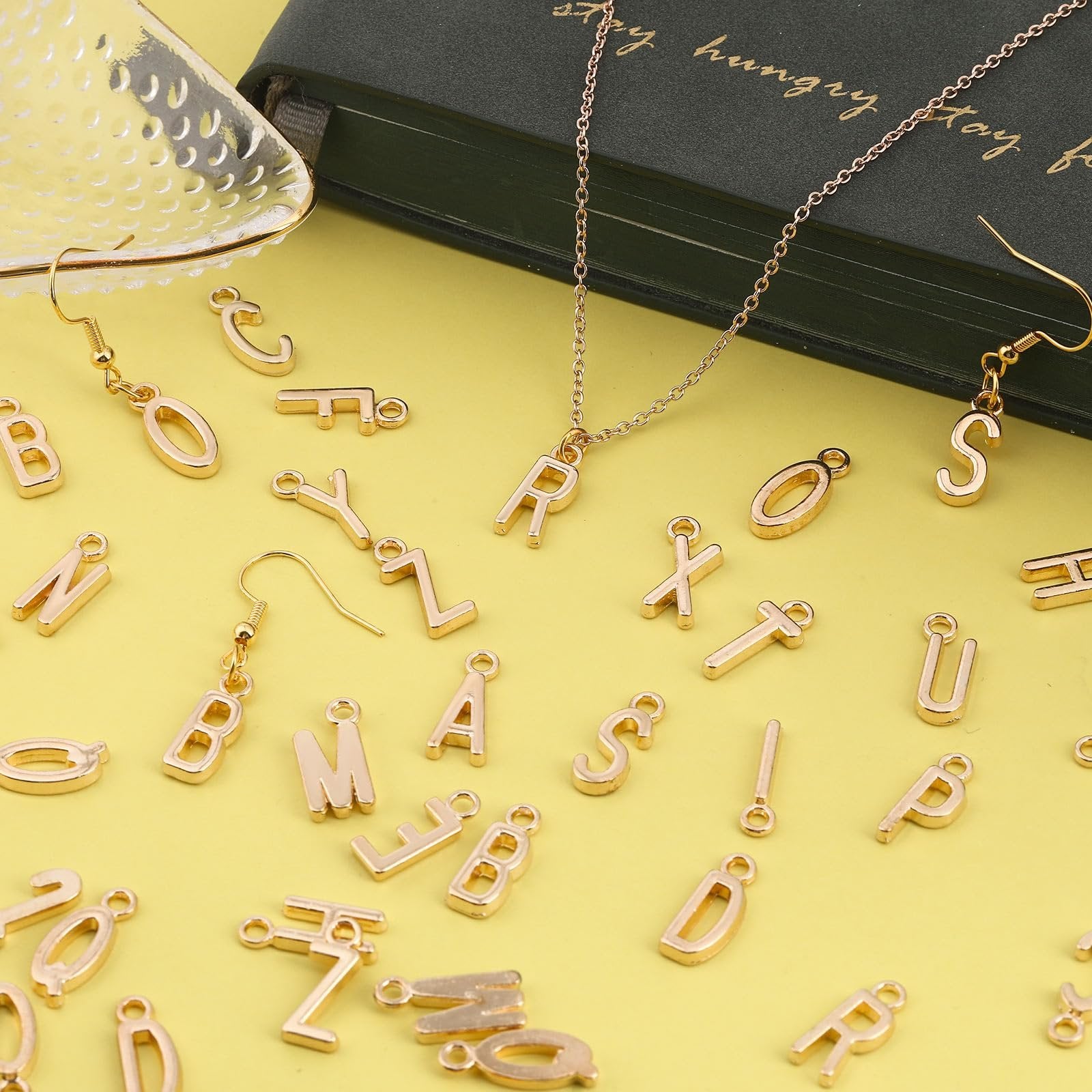 Gold alphabet letter charms and earrings scattered on a yellow background with a green book and decorative tray.