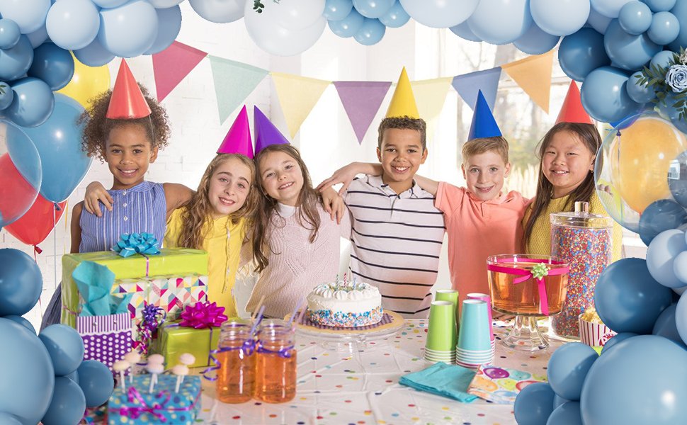 Six smiling children in party hats gathered around a table with gifts and a cake, surrounded by balloons.