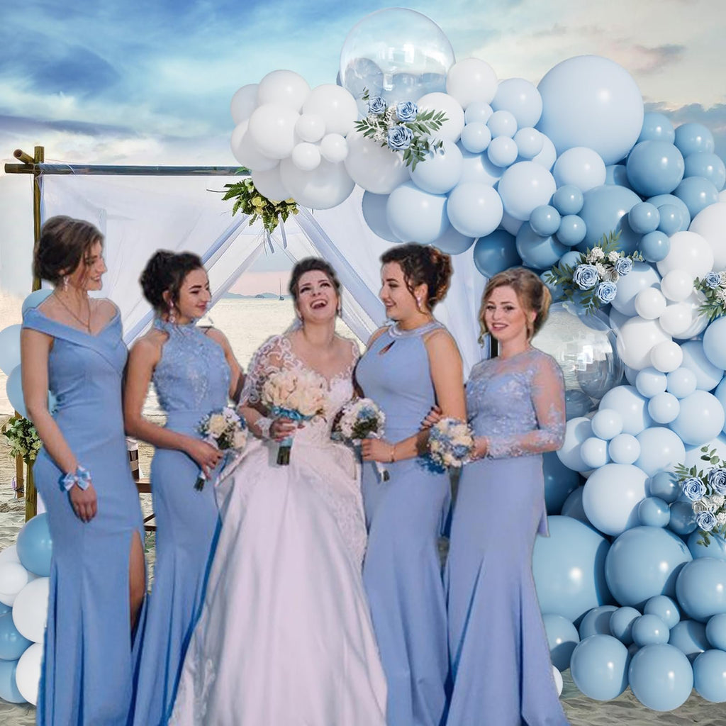 Bride and bridesmaids in blue dresses posing in front of a dusty blue and white balloon arch at a beach wedding.